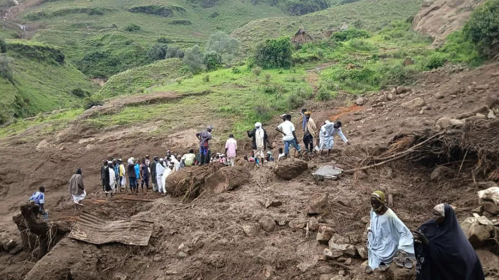 Survivors and rescue workers gather amid debris in Darfur after a deadly landslide, with damaged homes and mud-covered terrain visible.